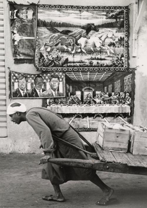 Man In The Street: a laborer hauls his load through the bazaar of Manama, Bahrain. The pictorial wall hangings above him are displayed for sale in one of the narrow streets of the Persian Gulf island