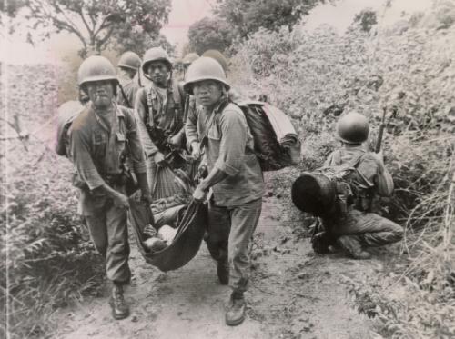 A platoon leader, seriously wounded when he stepped on a mine, is carried by Vietnamese soldiers retreating from the mountain where they had attacked the Viet Cong unsuccessfully. Along the side of the road, other soldiers watch for snipers in the thick growth