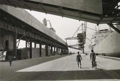 Street scene with man riding alongside boat at shipping dock, Guinea