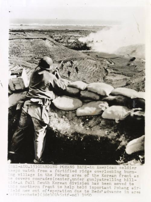 Guarding Pohang Base: an American soldier keeps watch from a fortified ridge overlooking burning village in the Pohang area of the Korean front as he covers comrades (center, under gun) patrolling hillside. A full South Korean division has been moved to this northern front to help hold important Pohang airfield now out of operation due to Reds' advance in area.
